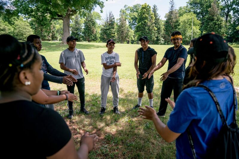 A group of young people stand in a circle on a grassy field, possibly in a park or outdoor setting. They appear to be engaged in some kind of activity or game, with their hands positioned in the center of the circle. Some are wearing casual clothing, while others have hats or headbands. The background includes trees and greenery, suggesting a natural environment.